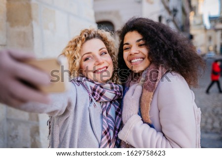 Outdoor close up portrait of two young beautiful fashionable happy smiling girls making selfie on street. City lifestyle. Ladies wearing stylish winter coats, beanie hats. Cristmas fair on background