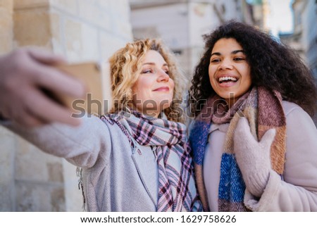 Two blond and brunette curly smiling sisters wth smartphone are taking selfie in the shopping time at winter holiday in the city center.