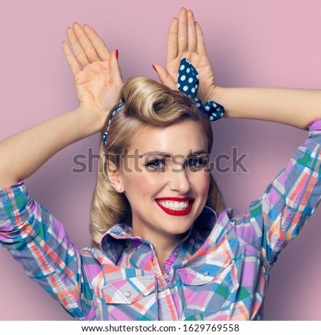 Funny bunny! Excited happy woman holding hands over head in rabbit ears gesture. Pin up girl in retro fashion vintage concept. Pink background. Square composition picture. 