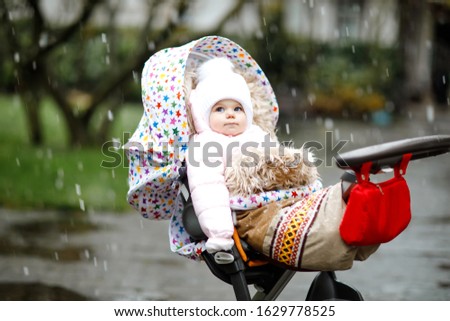Cute little beautiful baby girl sitting in the pram or stroller on cold day with sleet, rain and snow. Happy smiling child in warm clothes, fashion stylish baby coat. Winter or autumn day.