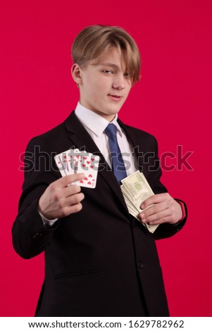 A teenager boy in a black jacket holds playing cards and a pack of dollars banknotes in his hands and poses on a red background