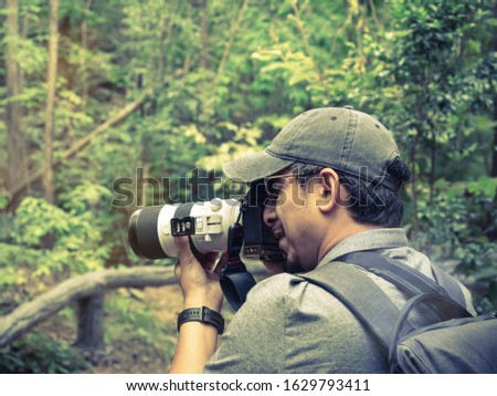 Asian Male backpacking Photographer wearing a black cap and glasses is shooting a beautiful landscape photo in the green forest. Color tone.