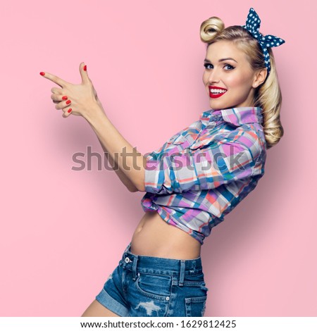 Square composition picture - woman pointing at something. Excited girl in pin up, showing some product or copy space for some text. Retro fashion and vintage. Pink background.