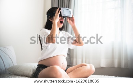 Asian pregnant women sitting in a white bed in the bedroom using a virtuar reality machine or VR, For relaxation to technology concept.