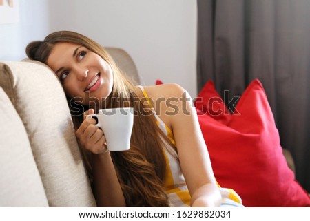 Young smiling woman sitting on sofa and looking up while drinking hot tea. Girl dreaming or thinking on couch at home in a leisure time.