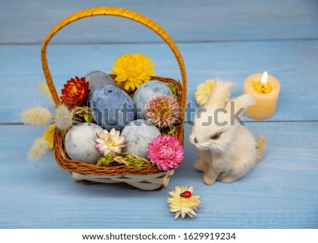 A basket full of Easter eggs painted blue, a hare, and burning candles on a wooden table.