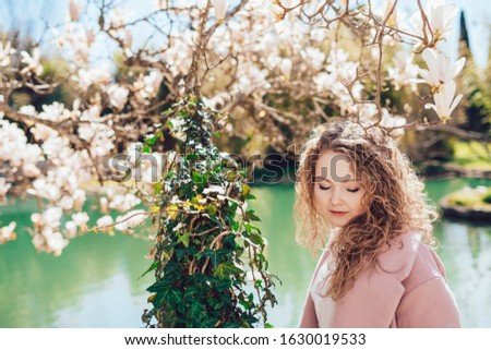 a girl with an angelic face sits downcast against a green pond above her head blooming magnolia branches