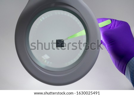 Hand in purple glove holding microchip with pair of tweezers under magnifying glass . Focus on chip.