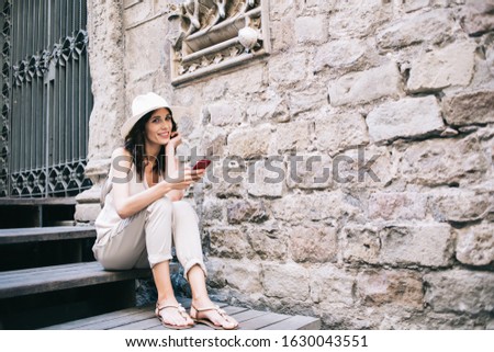 Happy young woman using smartphone and sharing photos with friends in social network while sitting on stairs next to historic building during exciting summer tour in city