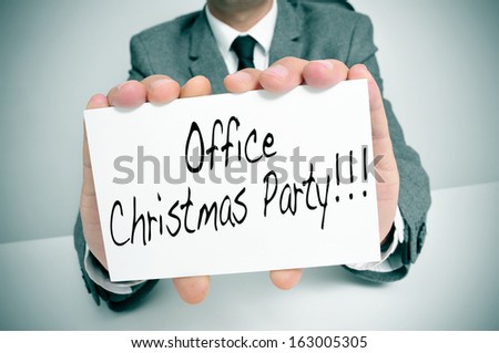a man wearing a suit sitting in a desk holding a signboard with the text office christmas party written in it