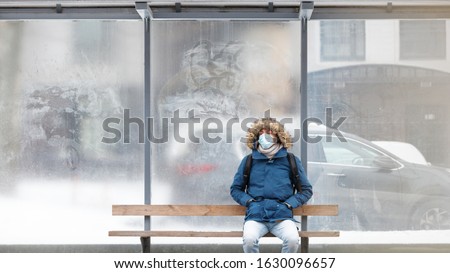 Sick man with a hood sitting alone on bench public transport, wearing protective facial mask against transmissible infectious diseases and as protection against the flu or coronavirus in public place Royalty-Free Stock Photo #1630096657