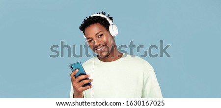 Young african american man listening to music from mobile phone using headphones over blue pastel background.