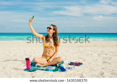Young woman in a swimsuit in the sand makes selfie on the beach on her smartphone