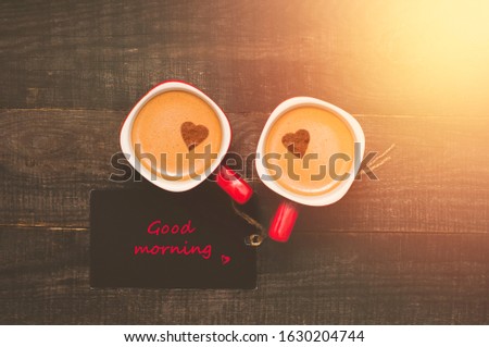 Red coffee cups of cappuccino or latte with hearts on a foam and a card note good morning on brown wooden background with sunlight. Top view. Flat lay