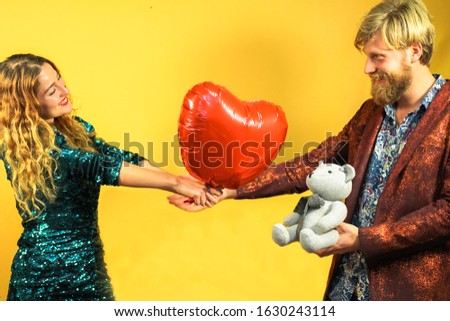 Man holding stuffed bear at San Valentin day. Woman holding red heart in his hands. Happy couple in a special day. Love concept. 
Yellow background. Focus hands girl and red heart - Image