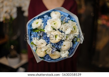 Woman holding a spring bouquet of tender white color roses and blue carnations in the wrapping paper in the blurred background of flower shop