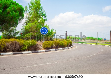 
Roundabout with road and roundabout sign with pine trees and bushes with blue sky