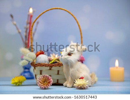 Horizontal photo of an Easter Bunny sitting at a basket of eggs, decorated with dried flowers. On a blurred background, a burning candle in a stand and willow branches.