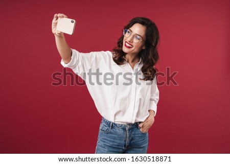 Picture of cheery optimistic happy young woman in glasses isolated over red wall background take a selfie by mobile phone.