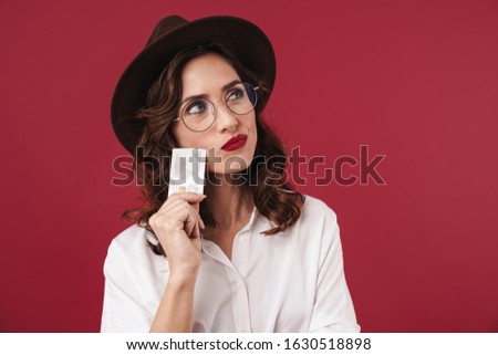 Picture of thinking thoughtful young woman in glasses isolated over red wall background holding credit card.