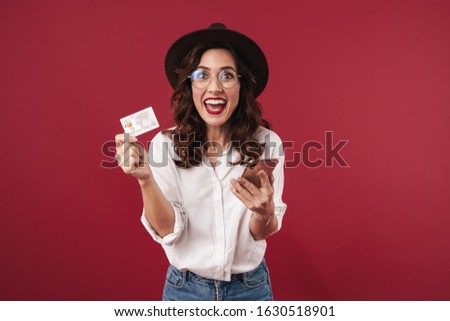 Picture of surprised optimistic young woman in glasses isolated over red wall background using mobile phone holding credit card.