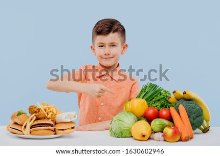 smiling little boy pointing at healthy food. fruits, vegetables and junk food. healthy food and fast food concept, isolated on blue background. studio. at the table Royalty-Free Stock Photo #1630602946