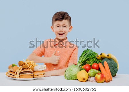 little boy showing thumb up with open mouth. vegetables, fruits or junk food. concept healthy food and fast food. isolated on blue background. studio. look in the camera