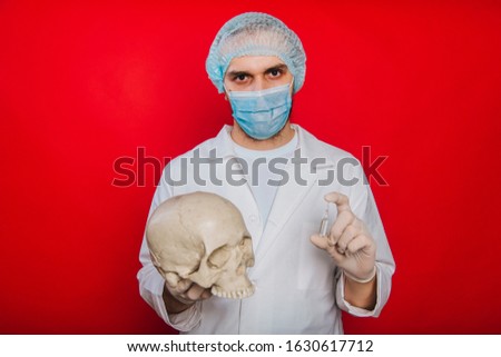 The doctor holds a skull and an ampoule with a vaccine in his hands. A young guy in a white coat, a medical mask and gloves on a red background. Pathologist medical worker. The concept of mortality fr