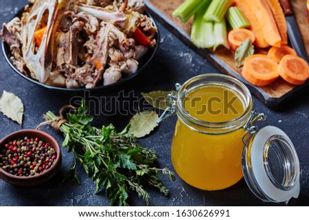 cooked chicken stock in a glass jar on a kitchen table with bouquet garni and chicken bones in a bowl, horizontal view from above