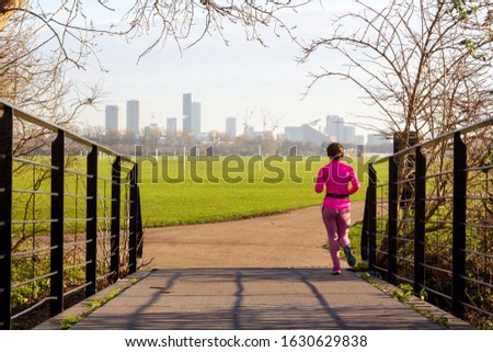 Young lady dressed in pink jogging in Hackney Marshes, a park in London, with the city skyline visible Royalty-Free Stock Photo #1630629838