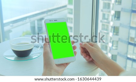 This stock photo features a woman holding her smartphone with green screen, sitting on the balcony of an apartment block with a morning coffee.