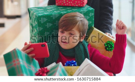 Close up boy sitting in shopping cart with Christmas present boxes in shopping mall use phone child family attractive teenager children cheerful happiness holiday kid smiling childhood slow motion