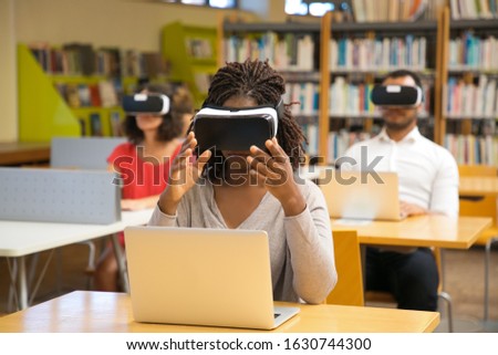 Concentrated young woman with virtual reality glasses. Focused woman sitting in library and experiencing new device. Technology, innovation concept