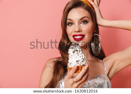 Picture of a young cheerful positive woman in bright sequins dress isolated over pink wall background holding cupcake wearing holiday birthday hat.