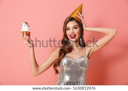 Picture of a young happy woman in bright sequins dress isolated over pink wall background holding cupcake wearing holiday birthday hat.