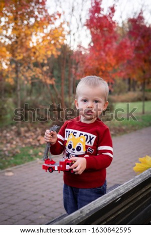 a blue-eyed fair-haired boy is standing in an autumn park by the bench, wearing a burgundy sweater with a picture of a fox and jeans, in his hands a fire truck