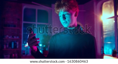 Smiling. Cinematic portrait of stylish redhair man in neon lighted interior. Toned like cinema effects in purple-blue. Caucasian model using smartphone in colorful lights indoors. Flyer.
