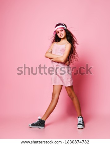 Young smiling slim teenager girl with long curly hair in translucent visor cap and pink striped dress is posing full body with her arms crossed on pink background