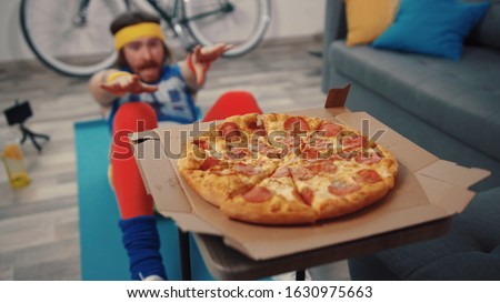 Funny fitness man doing abdominal crunches exercise failing to reach tasty hot pizza in the living room at home. Royalty-Free Stock Photo #1630975663