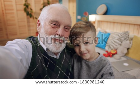Footage of granddad with gray hair and little boy taking selfie. Modern grandpa using camera, hugging little boy. Men smiling in background of bed. Bedroom.