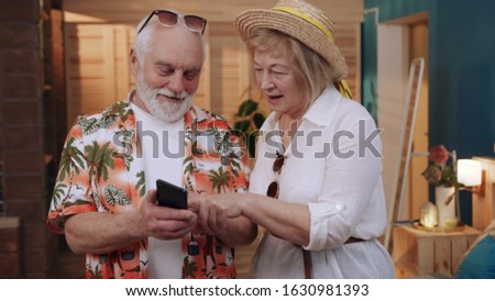 Old smiling couple in love holding smartphone in hands, reviewing photos in smartphone. Resort. Indoors. Elderly man and woman in casual clothes using gadget. Relax.
