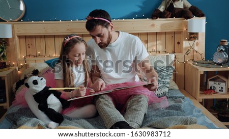 Beauty affectionate family portrait. Close-up zoom in view happy handsome dad and his daughter dressed like fairies reading a book together while sitting in bedroom. Home party.