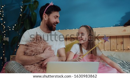 Cheerful pretty dad and daughter doing magic tricks with wand while watching cartoons on laptop. Family portrait of young man and his kid girl dressed like fairies sitting on bed at home.