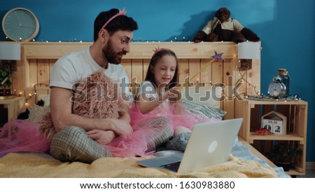 Fairytale family party at home. View of happy father and daughter playing fairies waving with magic wand looking on laptop screen sitting on bed and having fun.