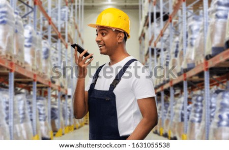 logistic business, technology and people concept - happy smiling indian loader or worker in helmet using voice command recorder on smartphone over warehouse background