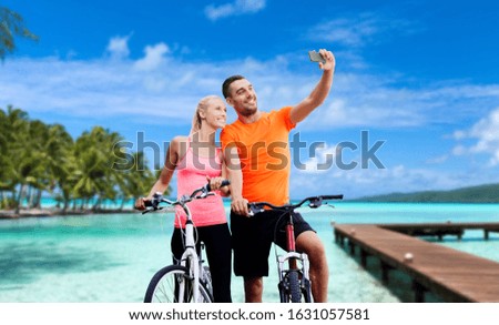 fitness, sport and people concept - happy couple with bicycles taking selfie by smartphone over wooden pier, palm trees and sea in french polynesia on background
