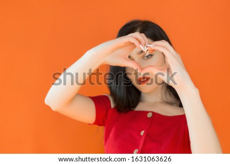a girl with black hair and a red dress, stands on an orange background, shows her hands with a heart