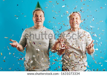 Horizontal studio portrait of young adult twin brothers celebrating their birthday with confetti,  blue background