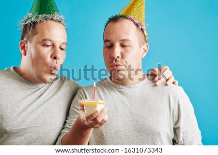 Horizontal studio portrait of young adult twin brothers standing together with small piece of birthday cake blowing out candle