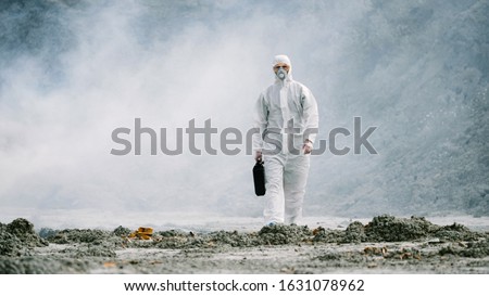 Lab technician in a mask and chemical protective suit, walks on dry ground with a tool box through toxic smoke.